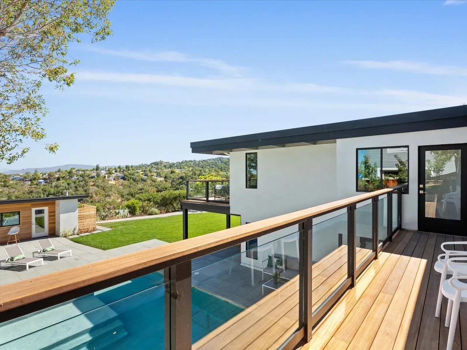Modern house with wooden deck, glass railing, and pool overlooking a green hillside.