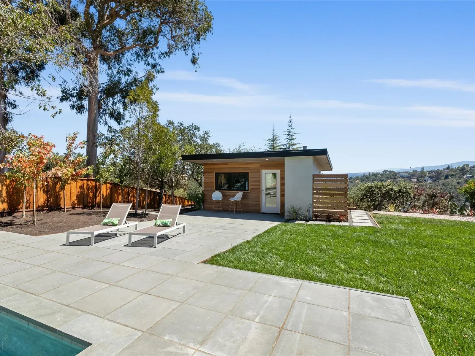 Backyard with modern shed, patio, lounge chairs, and green lawn under a blue sky.