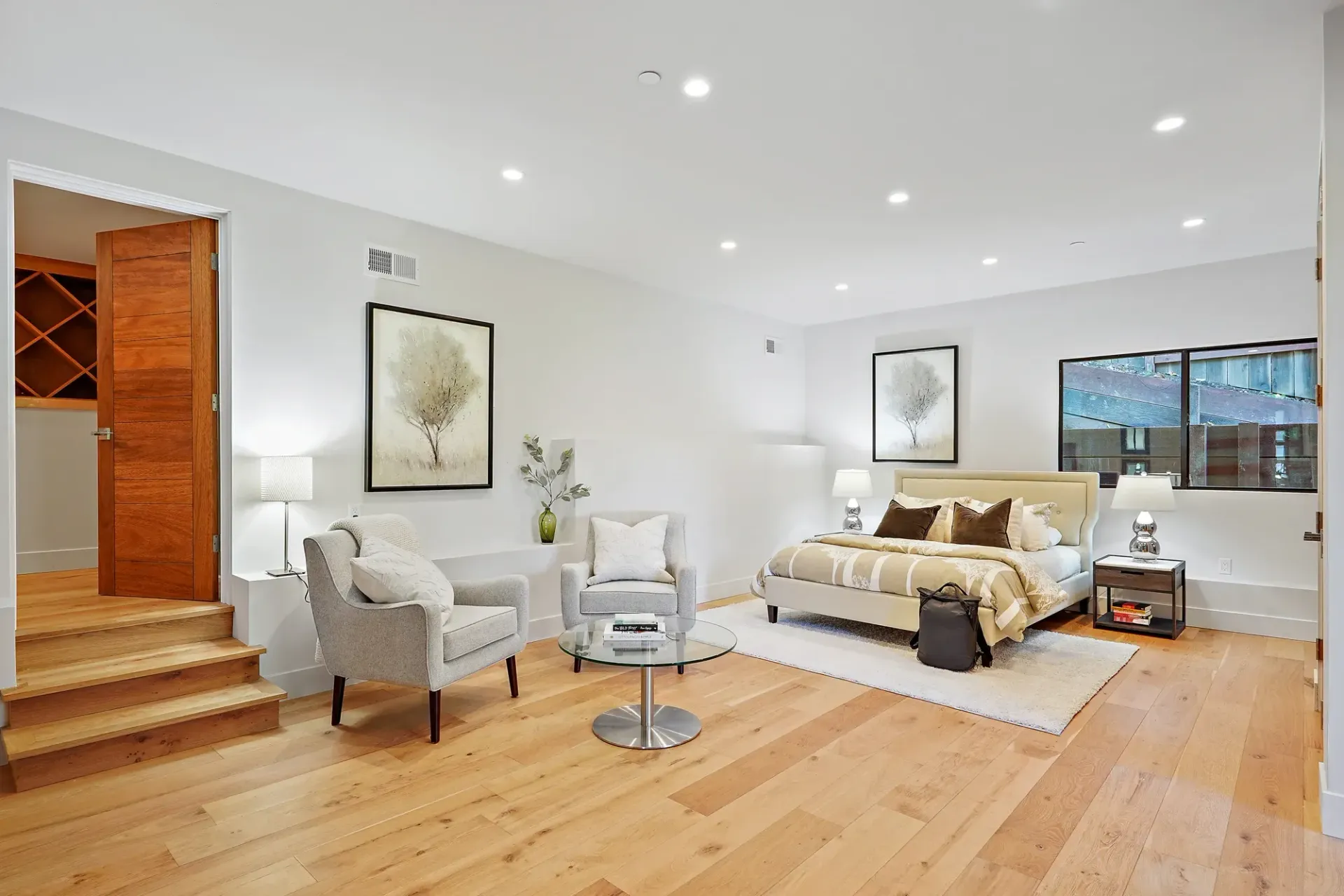 Bedroom with hardwood floors, two armchairs, bed, and artwork on white walls.