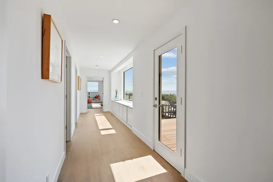 Long hallway with white walls, light wood floor, and a door and window with an outdoor view.