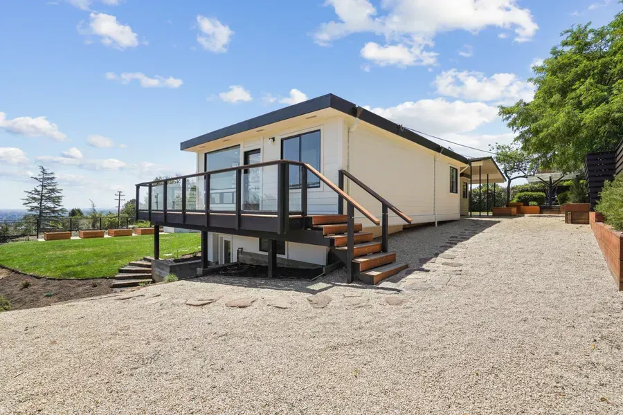 White house with black trim, deck, and gravel driveway on a sunny day.