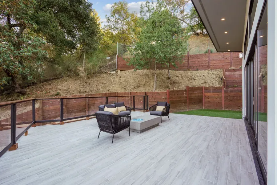 Outdoor patio with seating, wood deck, and view of hillside with trees and fence.