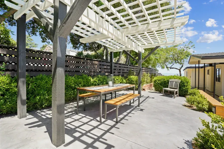 Outdoor patio with white pergola, wooden table and benches, surrounded by greenery.