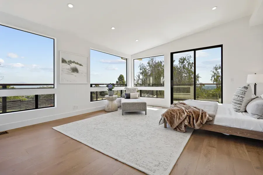 Bright bedroom with large windows overlooking a view. White walls, wood floor, rug, and bed.