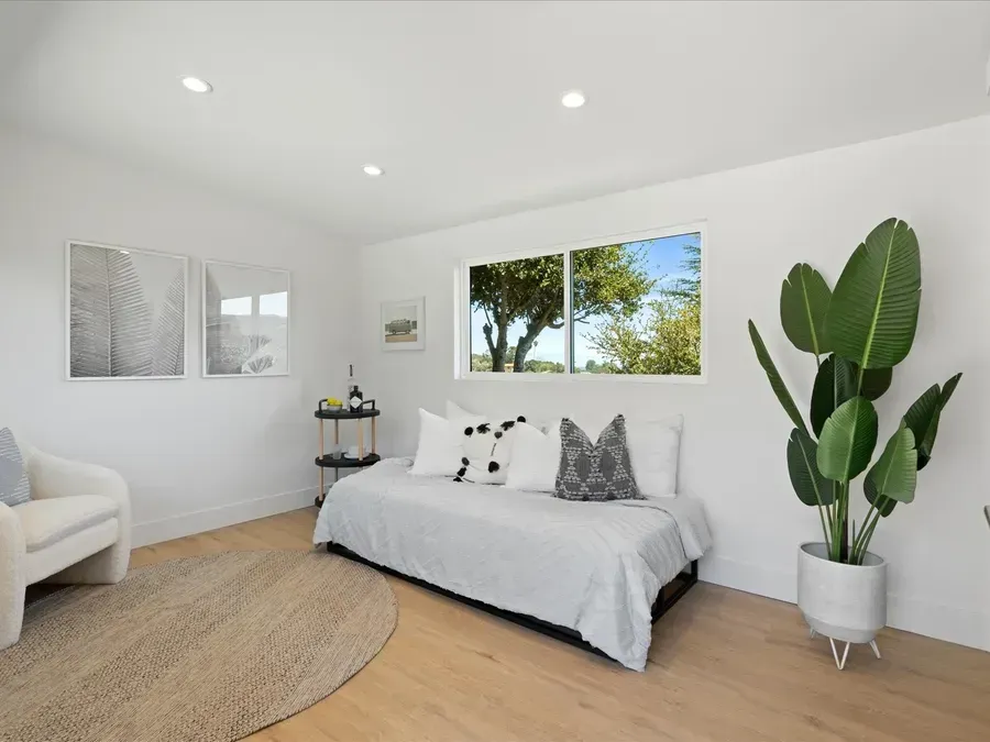 Bright room with a daybed, potted plant, and a window overlooking greenery. White walls and wooden floor.