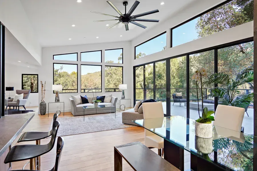 Bright modern living space with large windows overlooking trees. Includes a dining area, seating, and a bar.