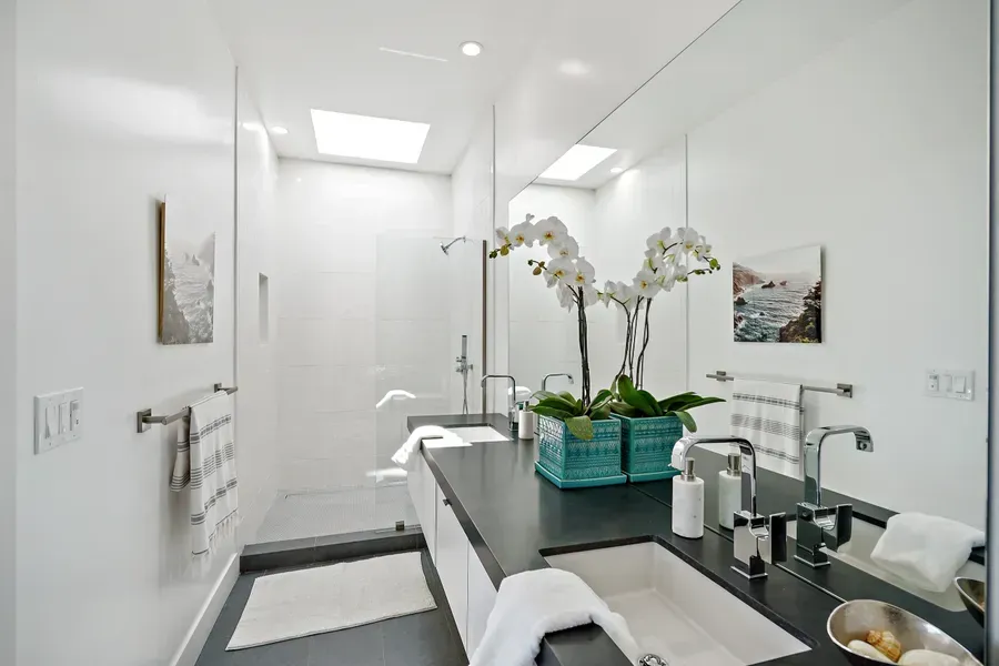 Modern white bathroom with a long dark countertop, large mirror, shower, and orchid plant.
