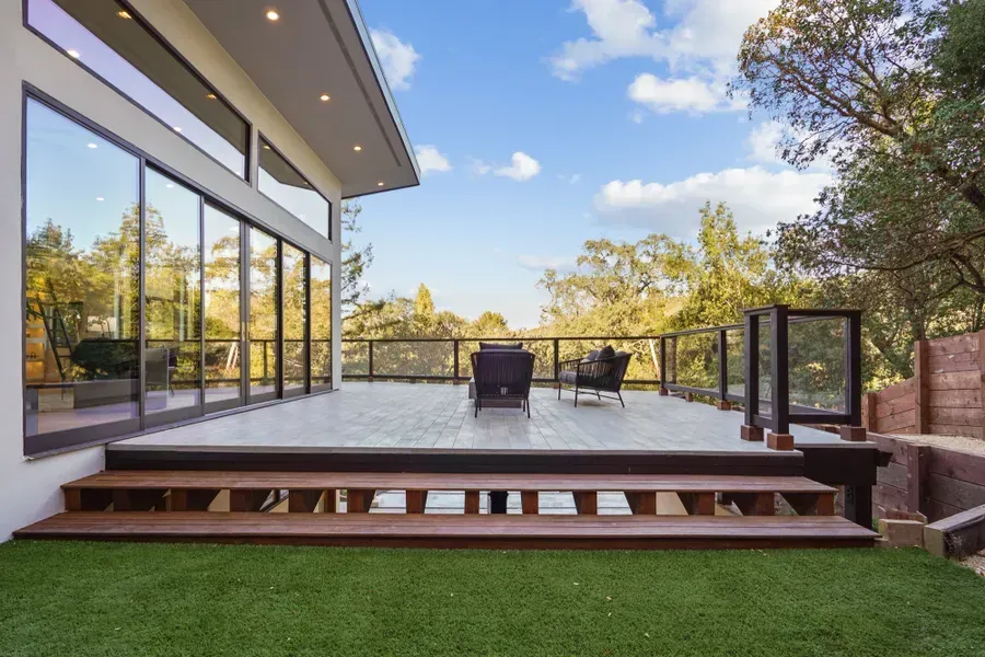 Modern house with glass doors opening to a wood deck, overlooking a tree-filled landscape.