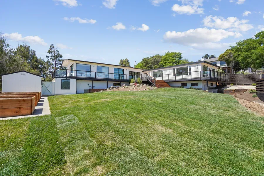 Backyard view of a white two-story house with green lawn and garden beds under a blue sky.