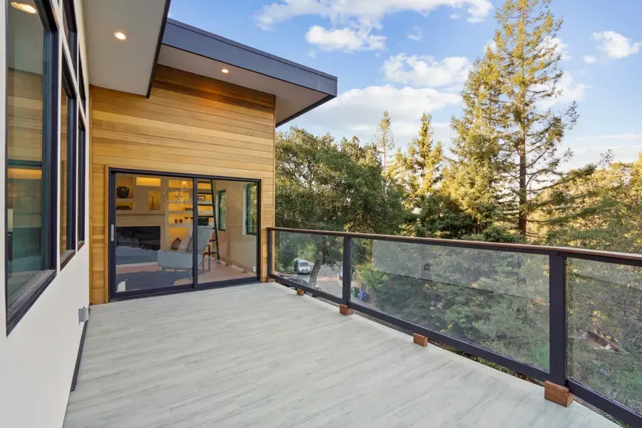 Balcony with glass railing overlooking trees, modern house exterior with wood siding and sliding glass door.