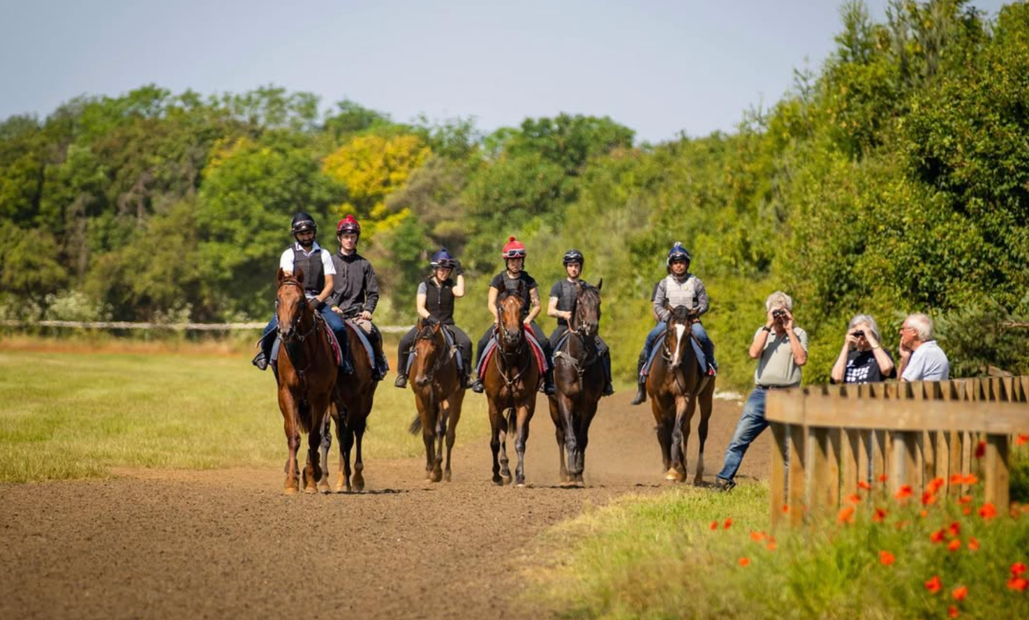 Group of riders on horseback on a dirt track, observed by two people. Trees in background, sunny day.