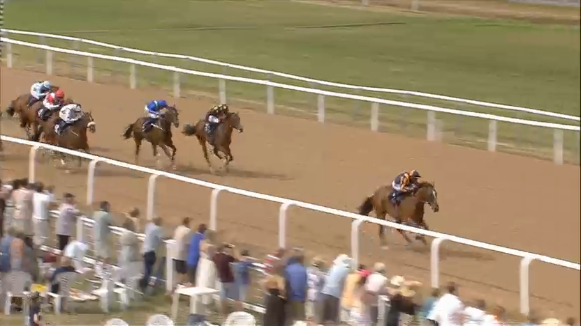 Horses racing on a dirt track with jockeys, spectators in the stands, a sunny outdoor setting.