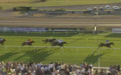 Horses racing on a track, jockeys riding. Spectators watch from the sidelines.