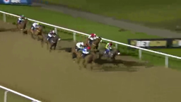 Horses racing on a dirt track at night, jockeys aboard, near a white fence.