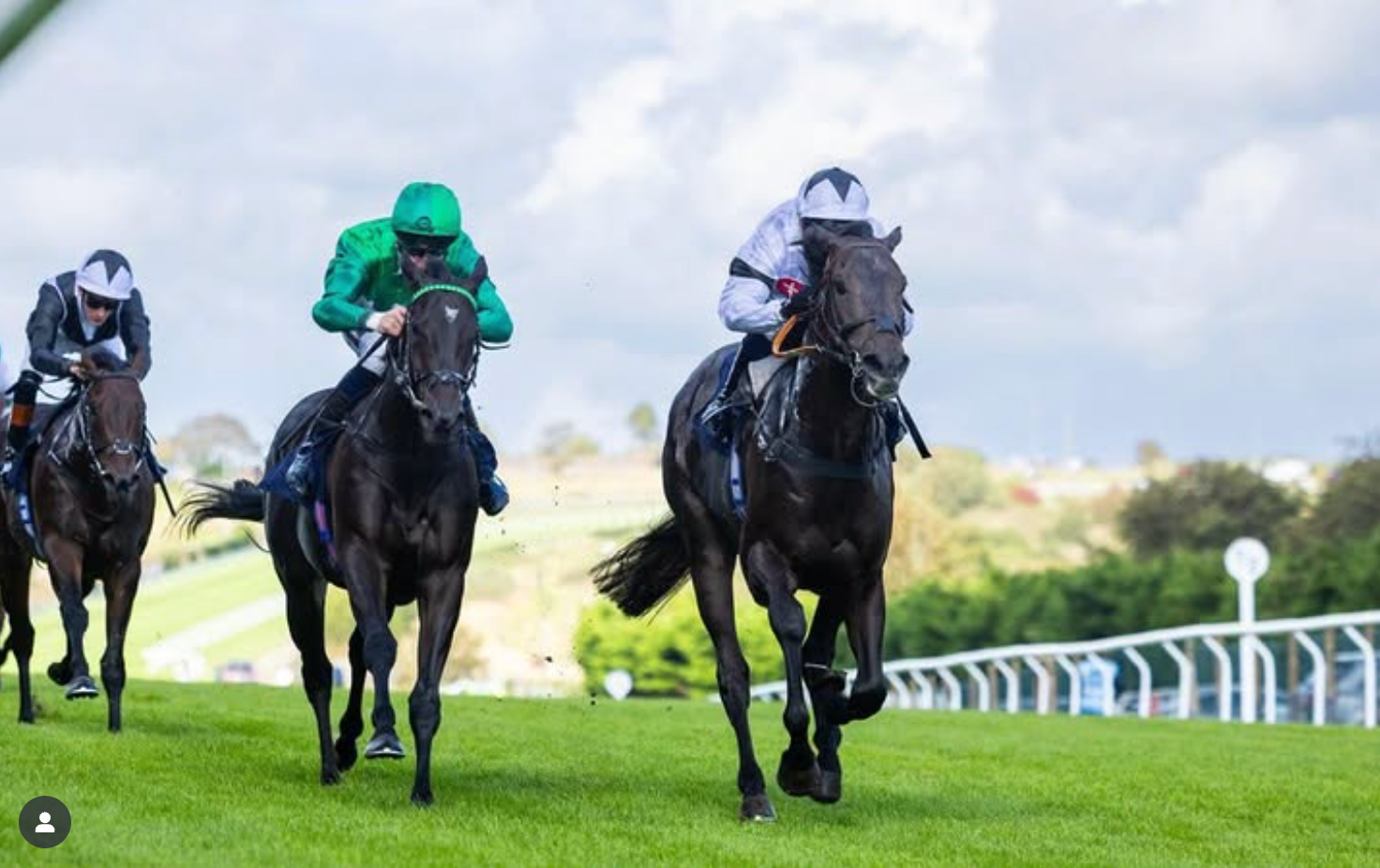 Horses racing on a green track, jockey in green leads the one with the white helmet in the foreground.