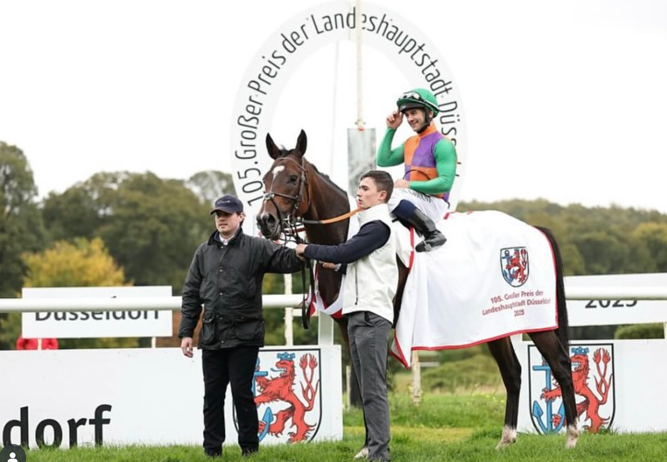 Horse and jockey on winner's platform with a person holding the horse. Text above says “Großer Preis der Landeshauptstadt Düsseldorf.”