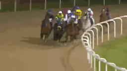Racehorses with jockeys rounding a turn on a dirt track at night.