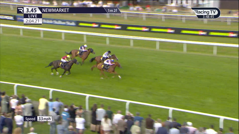 Horses racing on a green track at Newmarket, with spectators in the background.
