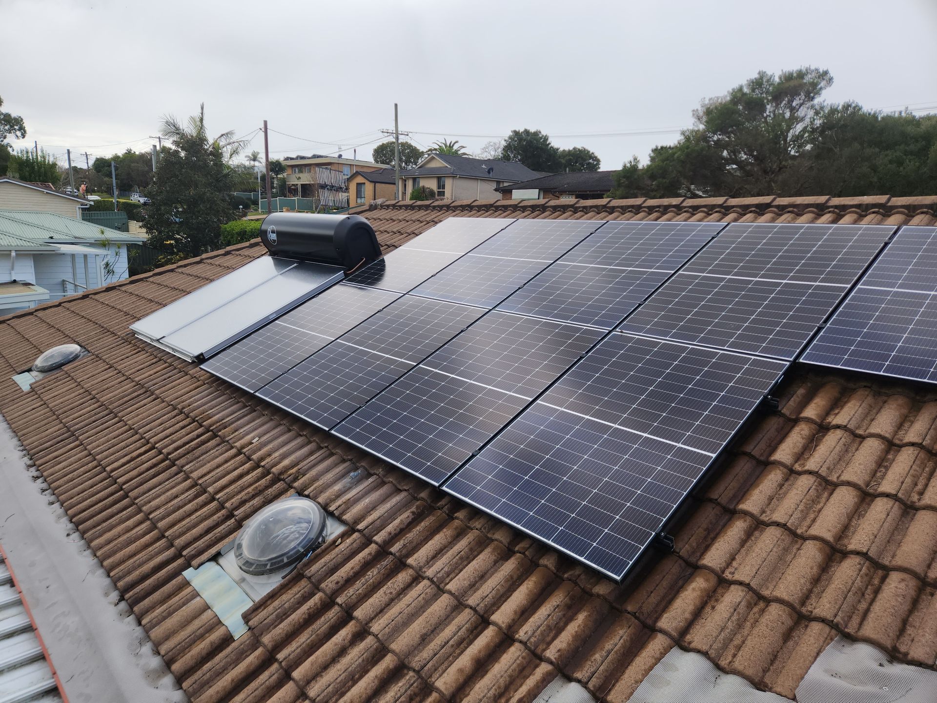 Solar panels and a black tank mounted on a terracotta-tiled roof under a cloudy sky — Solar Services in Erina, NSW