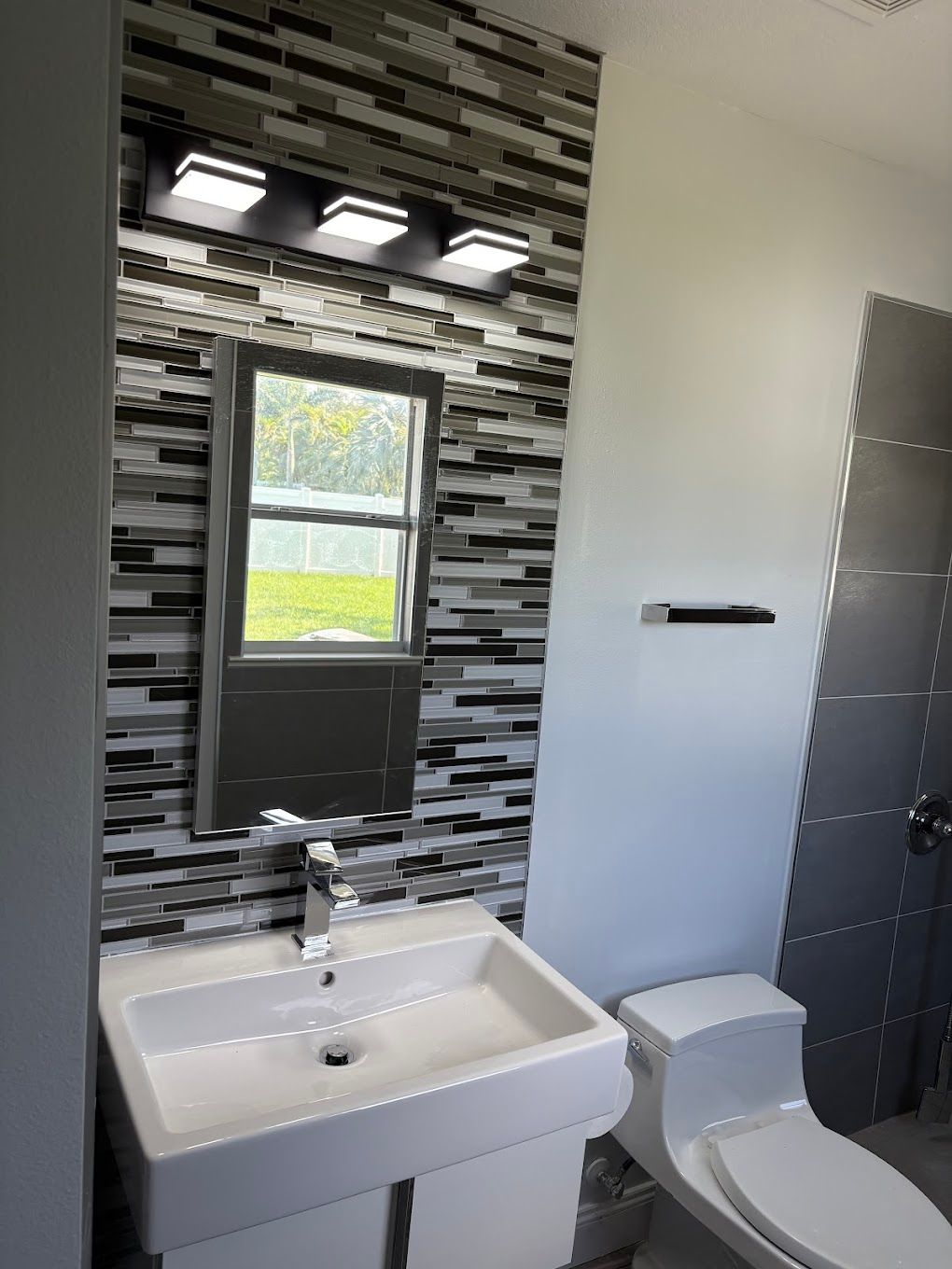 Modern bathroom with mosaic tile accent wall, vanity, toilet, and window.