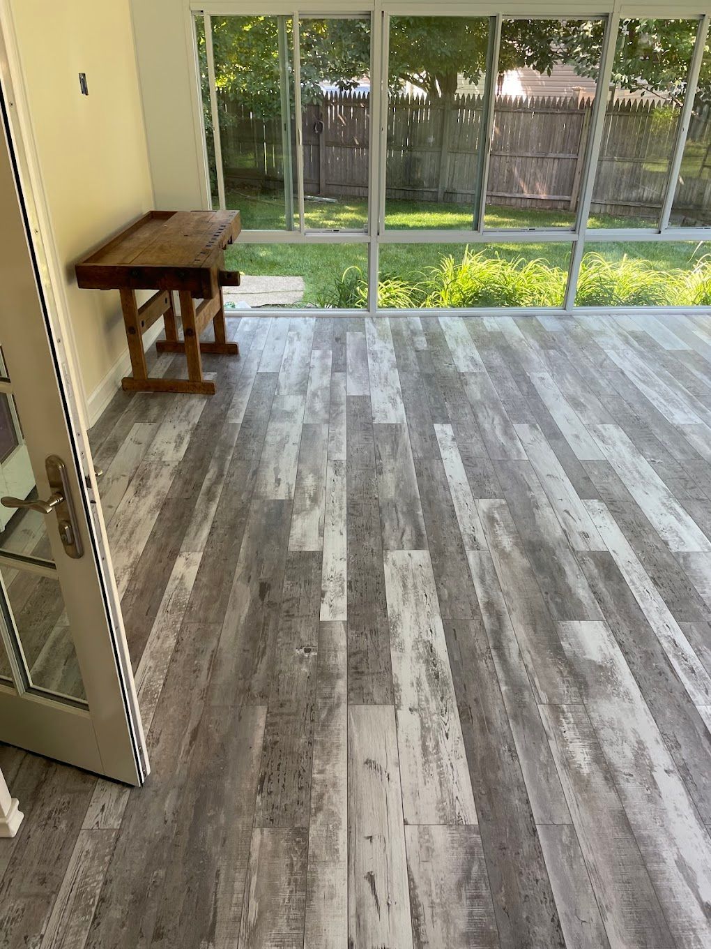 Sunroom with gray and white wood-look flooring, glass walls, and a wooden table; outside is a green yard.