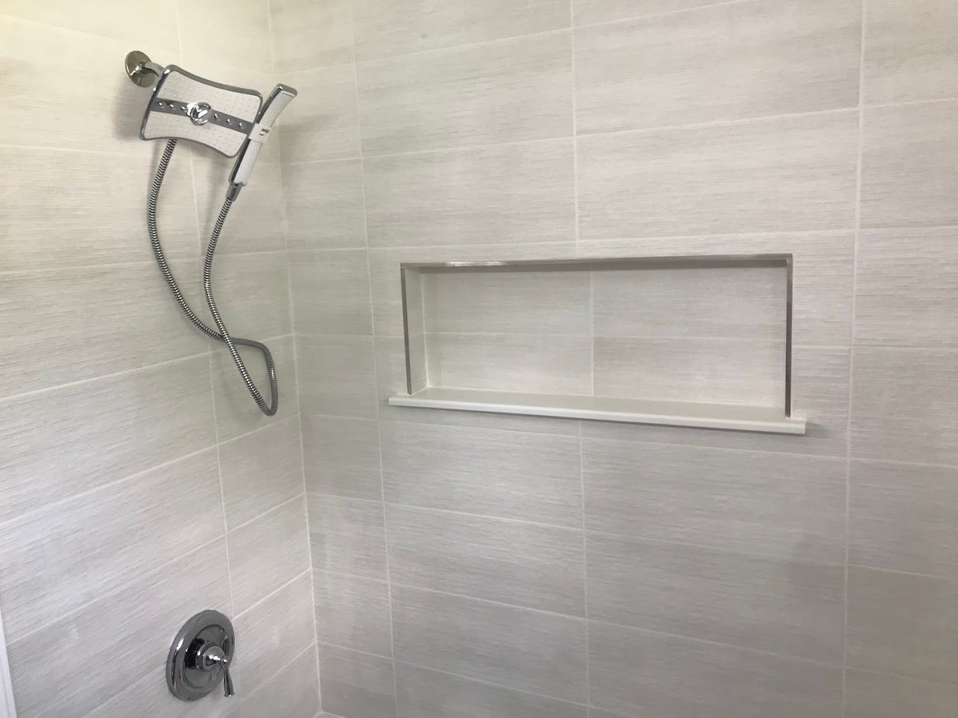 Shower with light gray tiles, chrome fixtures, and built-in shelf.