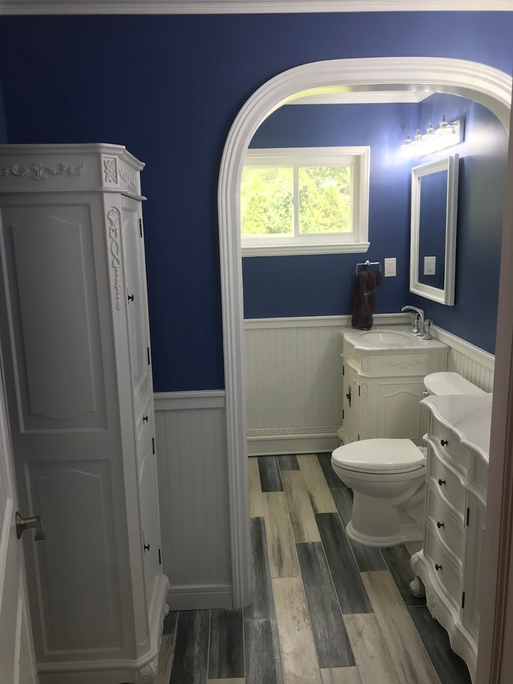 Bathroom with blue walls, white trim, and gray wood-look flooring. White vanity, toilet, and storage cabinet.