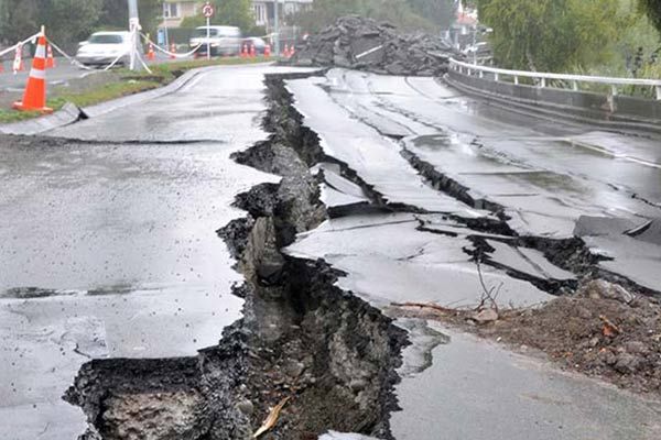 Road split by large earthquake crack and damage, with debris and orange cones on a wet street