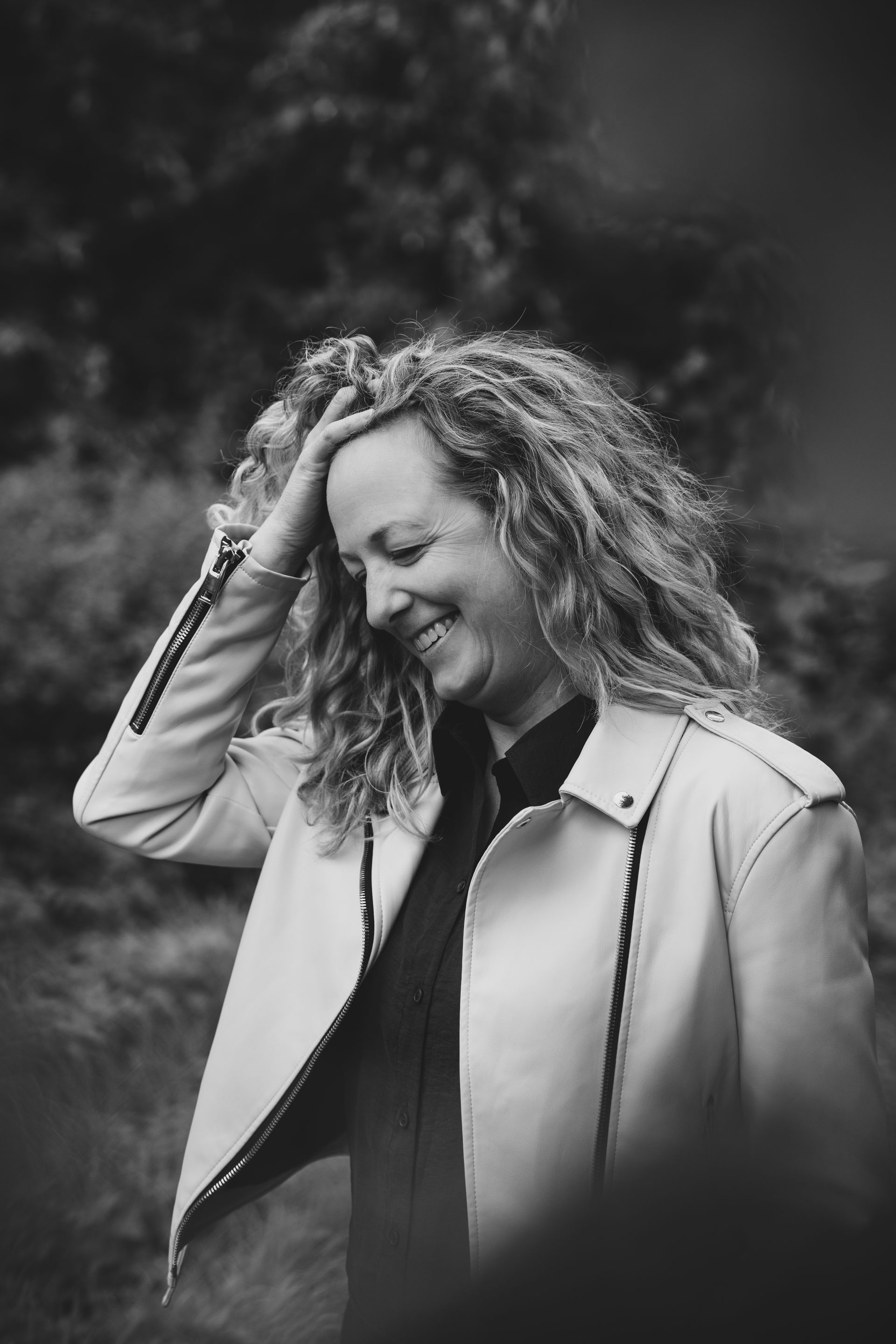 Vrouw met krullend haar glimlacht, met haar hand in haar haar, en draagt een witte jas. De achtergrond is wazig.