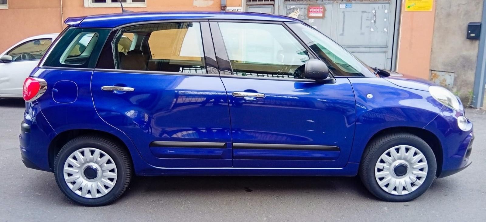 Fiat 500L blu parcheggiata in strada. Ha i coprimozzi bianchi. L'ambientazione sembra essere una città.