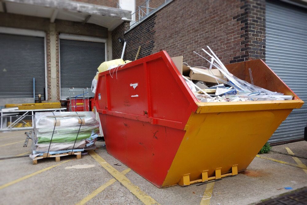 Red and Yellow Skip Bin Overflowing With Waste, Next to Building With a Roller Shutter Door — Hasti Skips in Bangalow, NSW