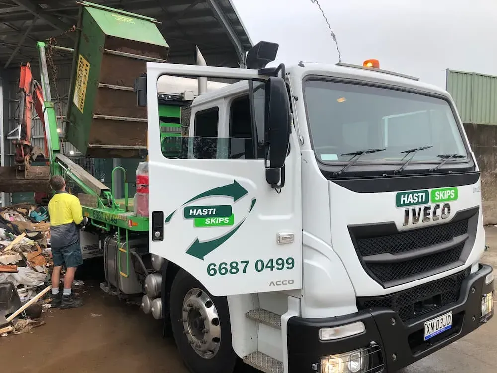 White and Green Truck Loading a Green Skip Bin at a Waste Site. Man Standing Next to It — Hasti Skips in Bangalow, NSW