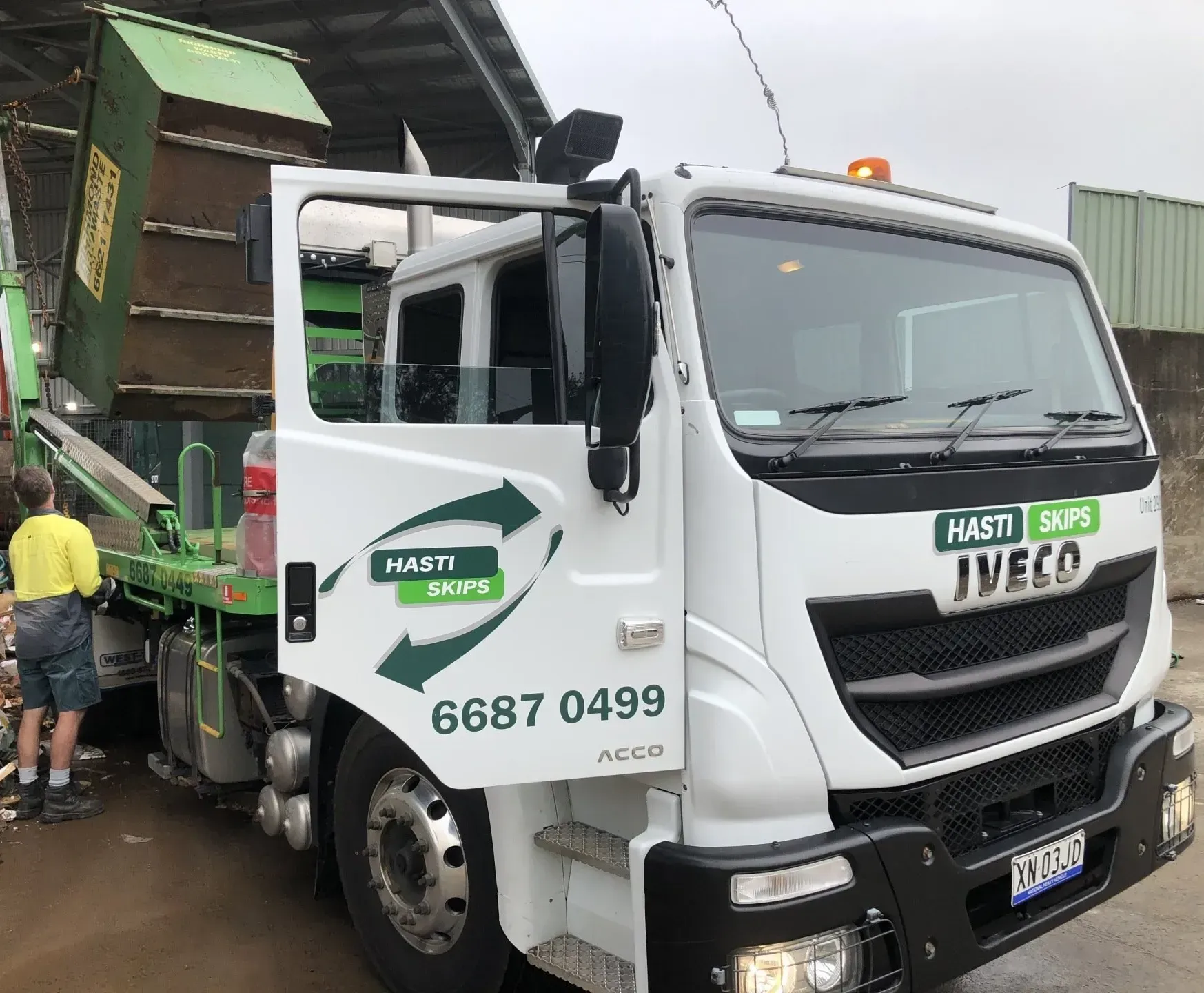 White Hasti Skip Truck With a Green Skip Being Loaded a Worker Stands Beside It — Hasti Skips in Casino, NSW