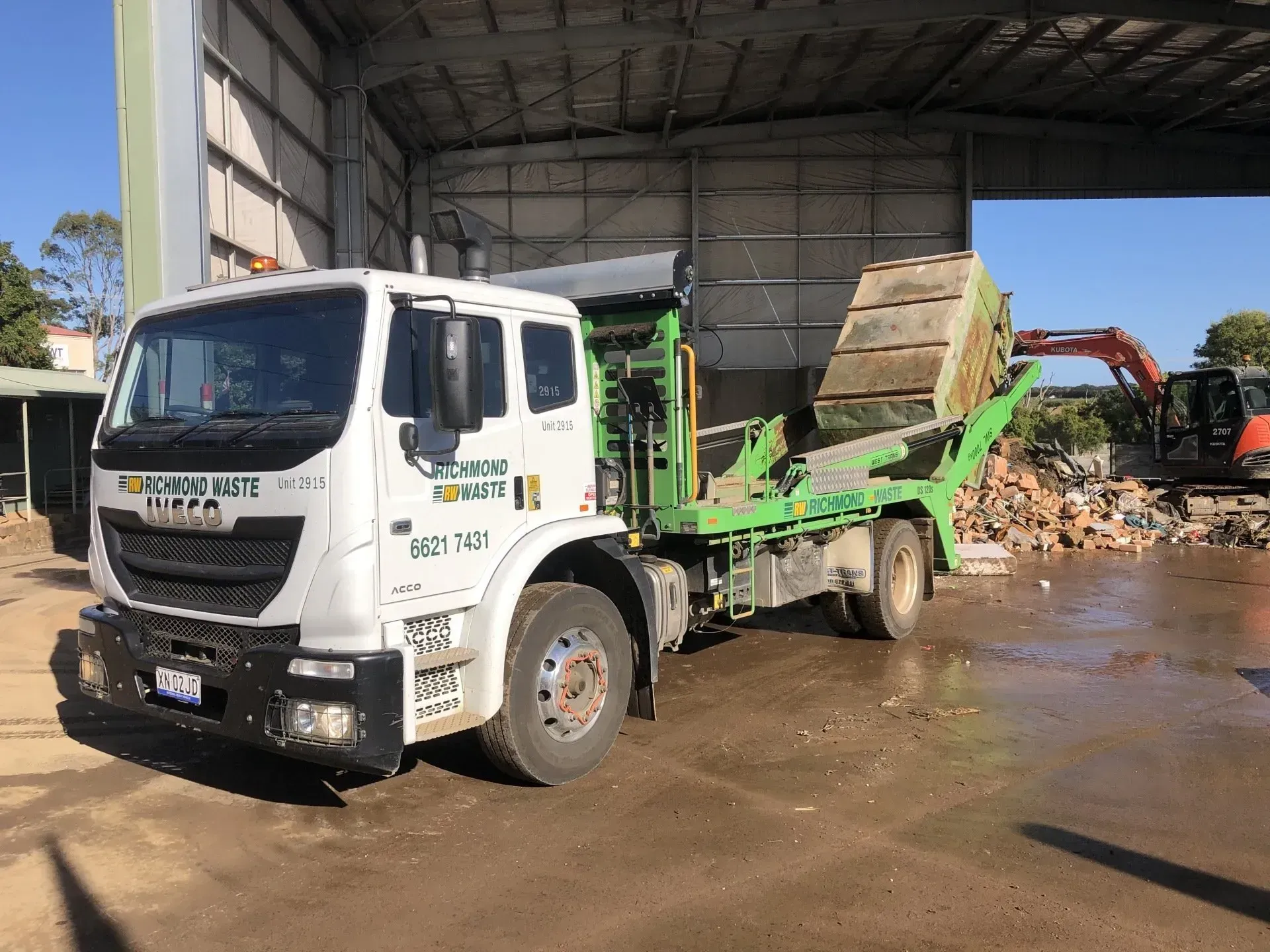 White Truck With Green Bed at a Recycling Center, Next to Debris Pile and Excavator — Hasti Skips in Bangalow, NSW