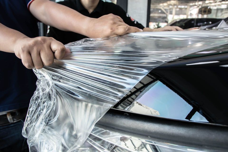 Hands stretch clear protective film over a black car panel in a garage.