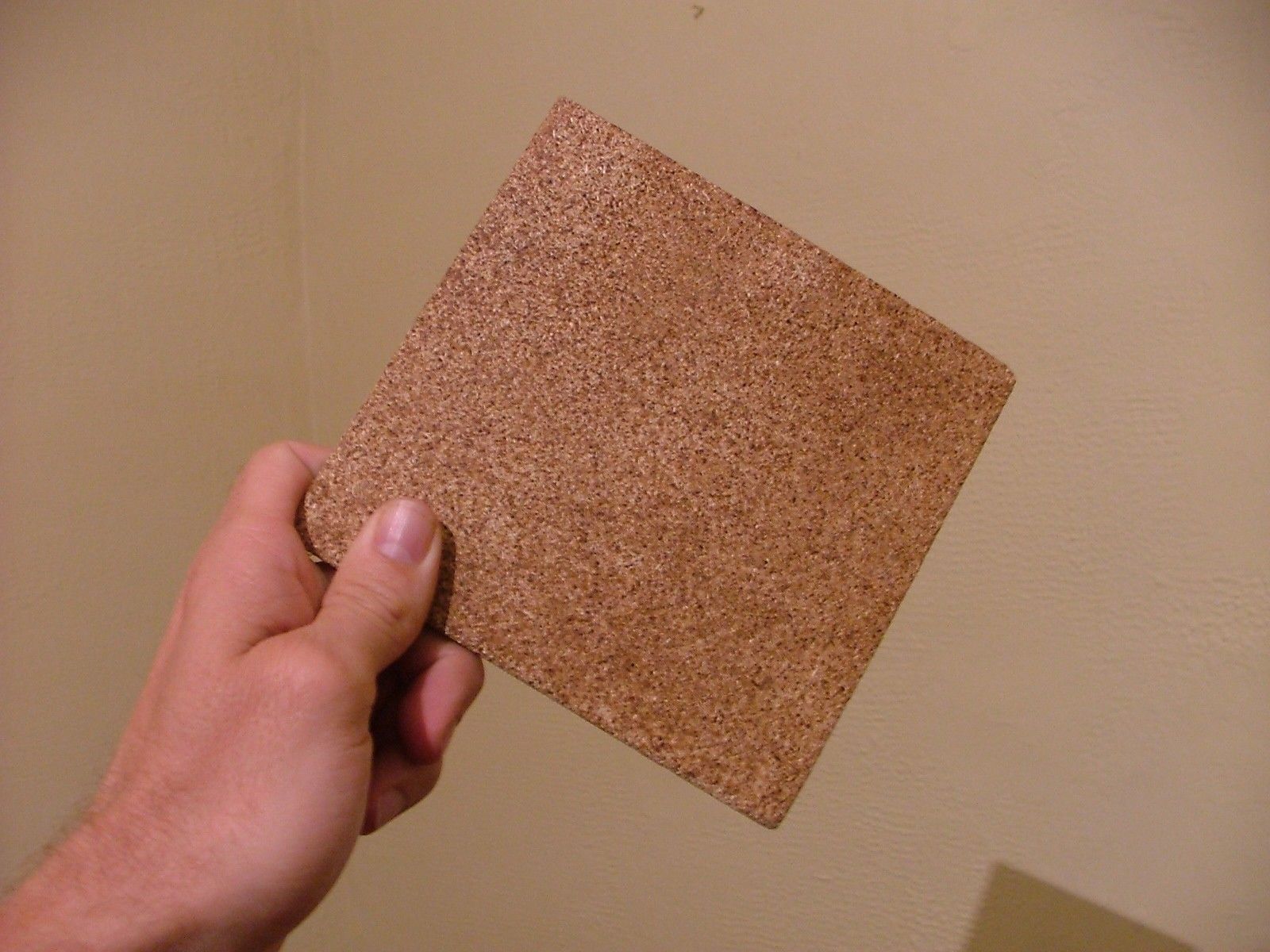 Hand holding a square cork tile, against a beige wall.