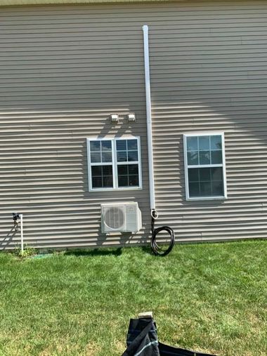House wall with two windows, an air conditioner, and a vertical pipe above a grassy yard.