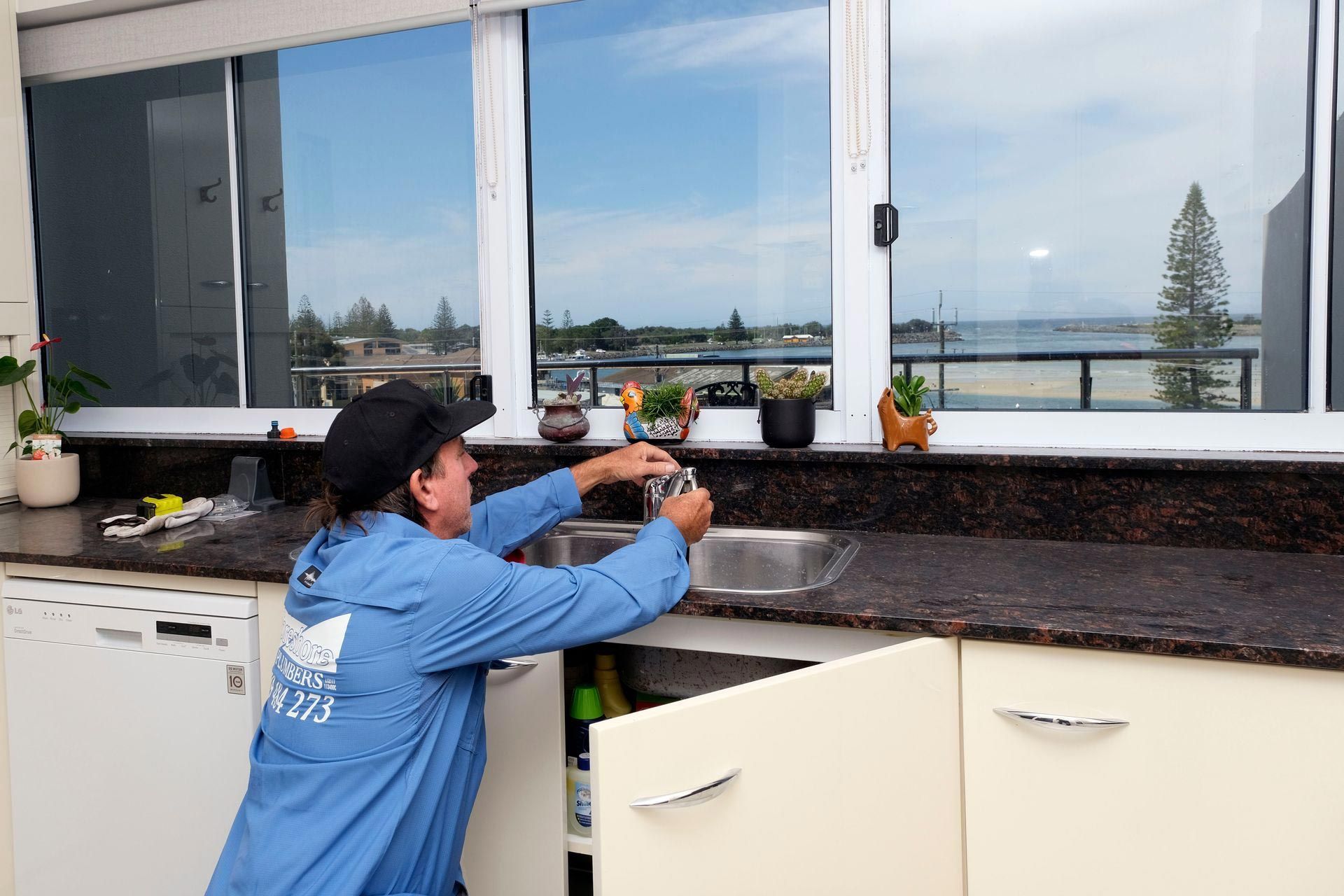 A Man in a Blue Jacket is Fixing a Sink in a Kitchen — Foreshore Plumbers In Tuncurry, NSW