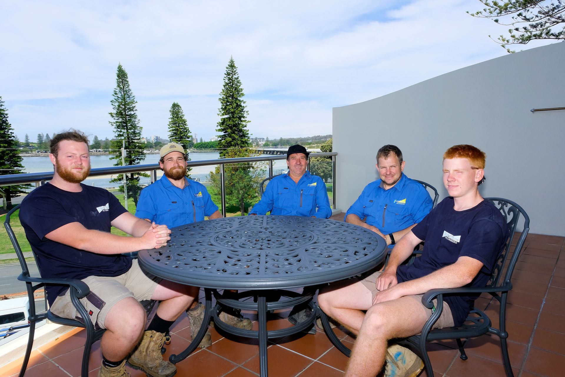 A Group of Men Are Sitting Around a Table on a Balcony — Foreshore Plumbers In Forster, NSW