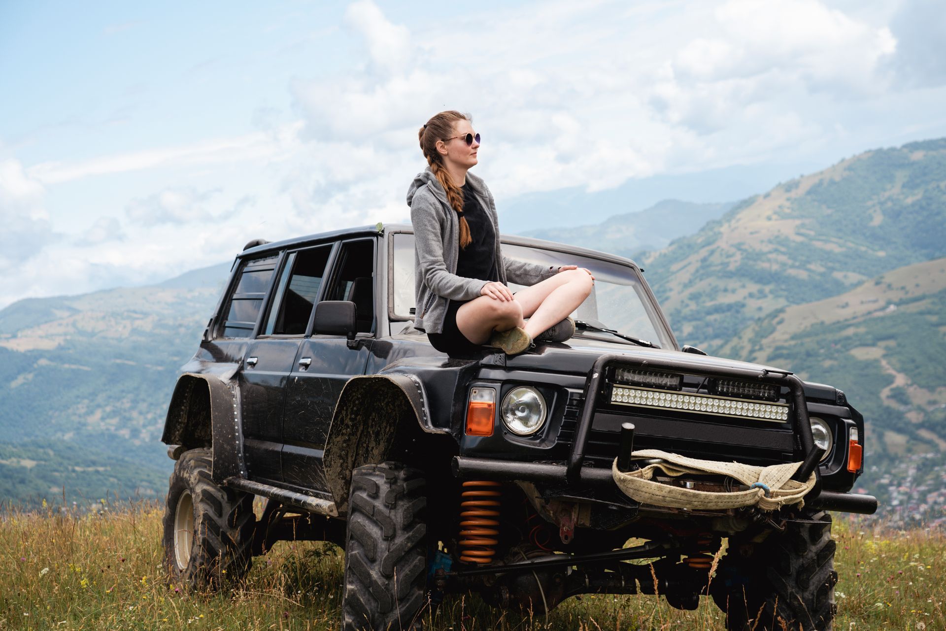 A girl sits on the hood of her off-road vehicle to look at the mountains.