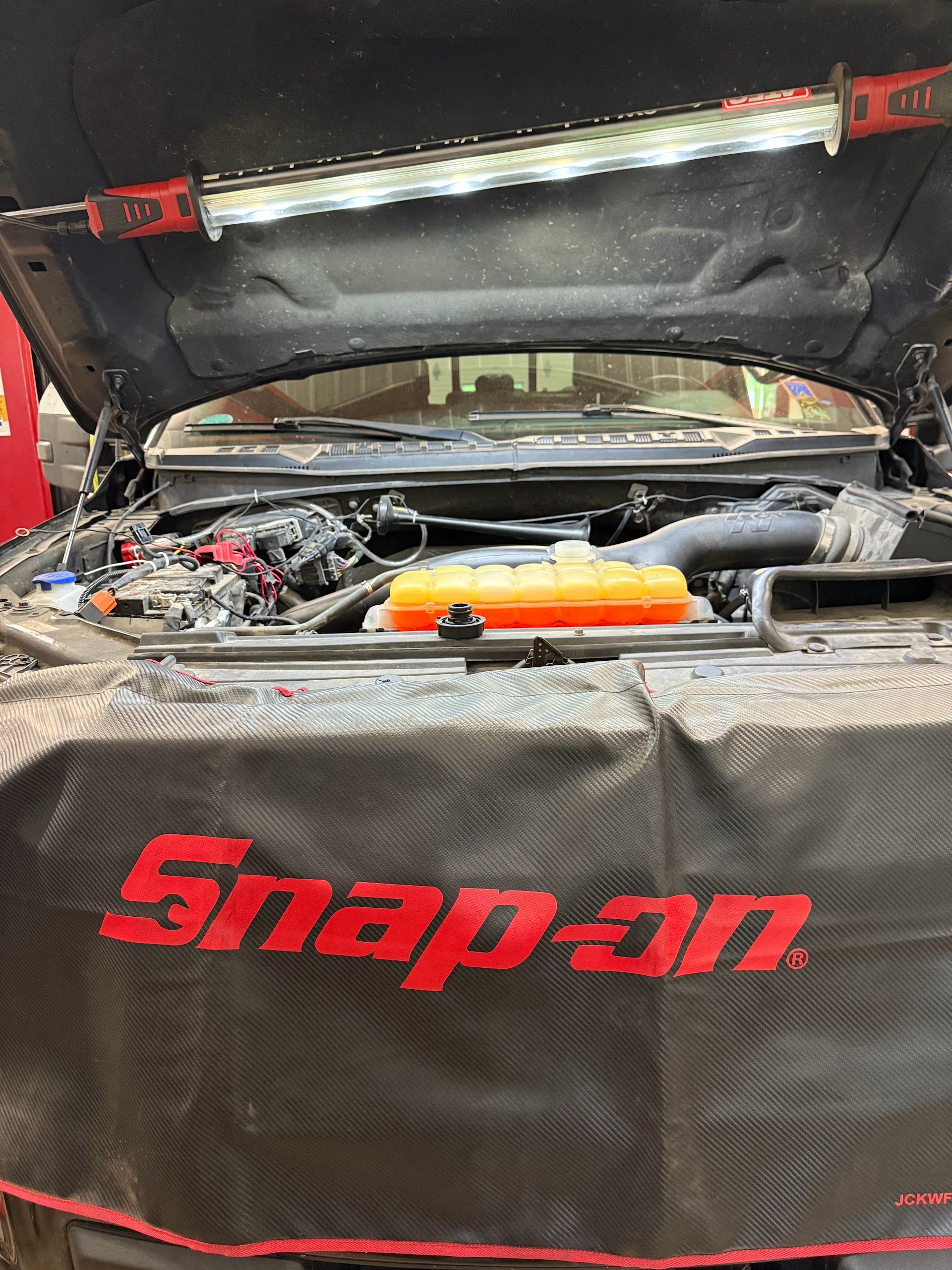 Two men smiling at the camera in a car repair shop with a car hood open in the background. | The Neighborhood Mechanic LLC