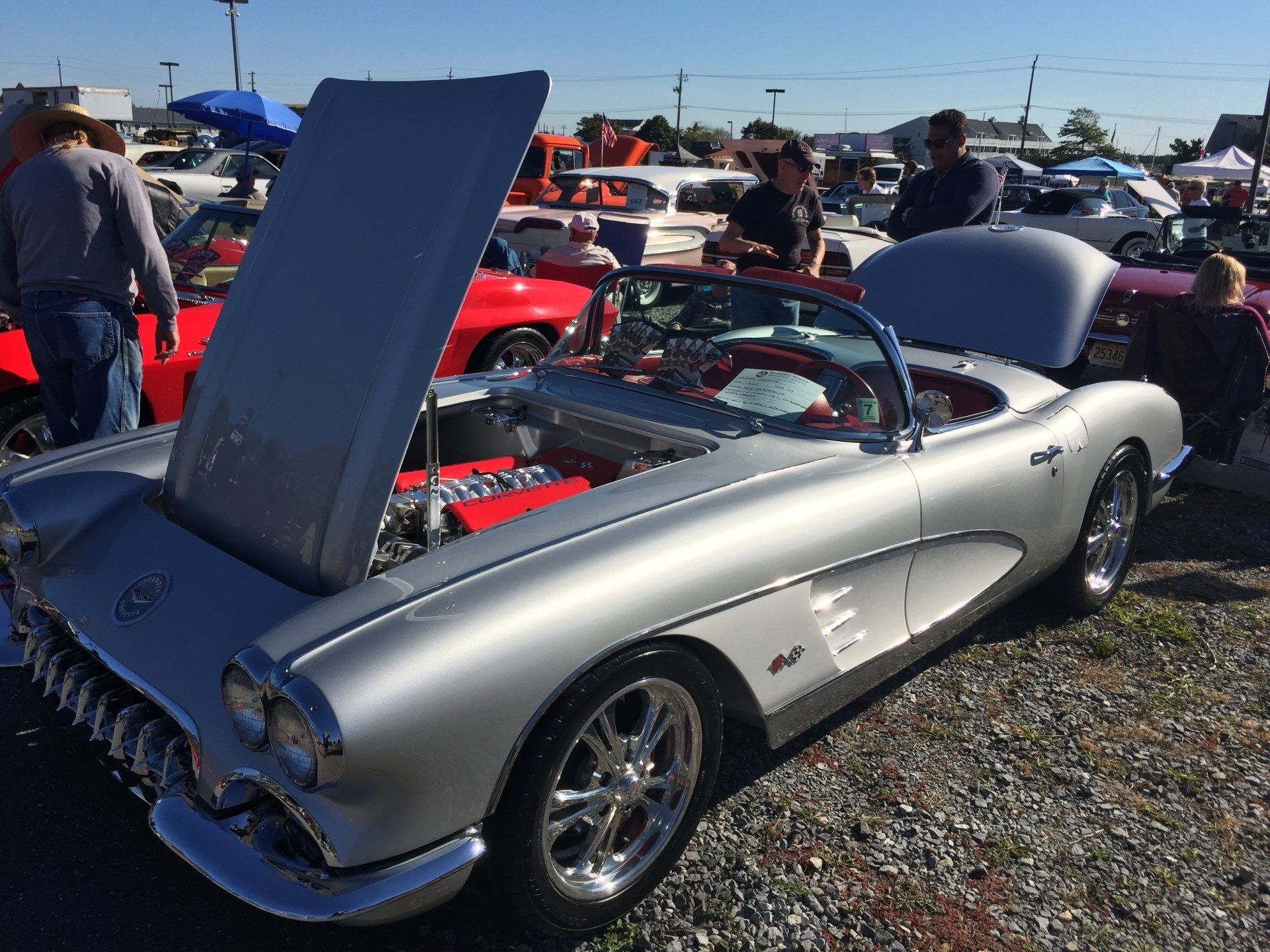 A silver car with the hood up is parked in a gravel lot