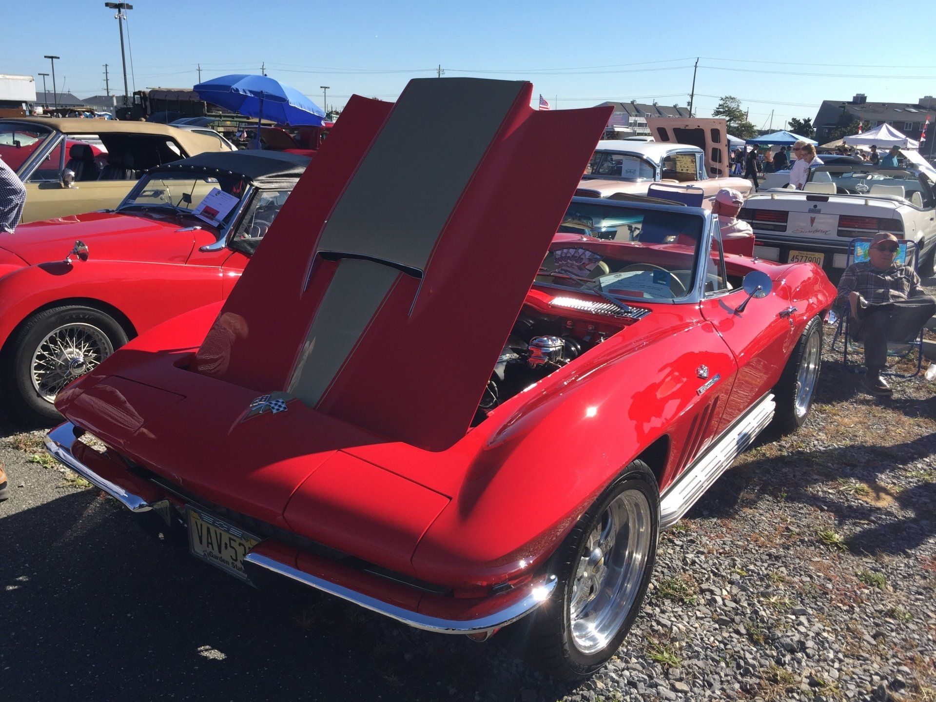 A red car with the hood up is parked in a gravel lot