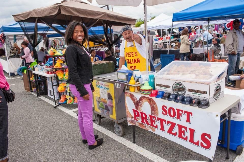 A woman is standing in front of a soft pretzel stand at a market.