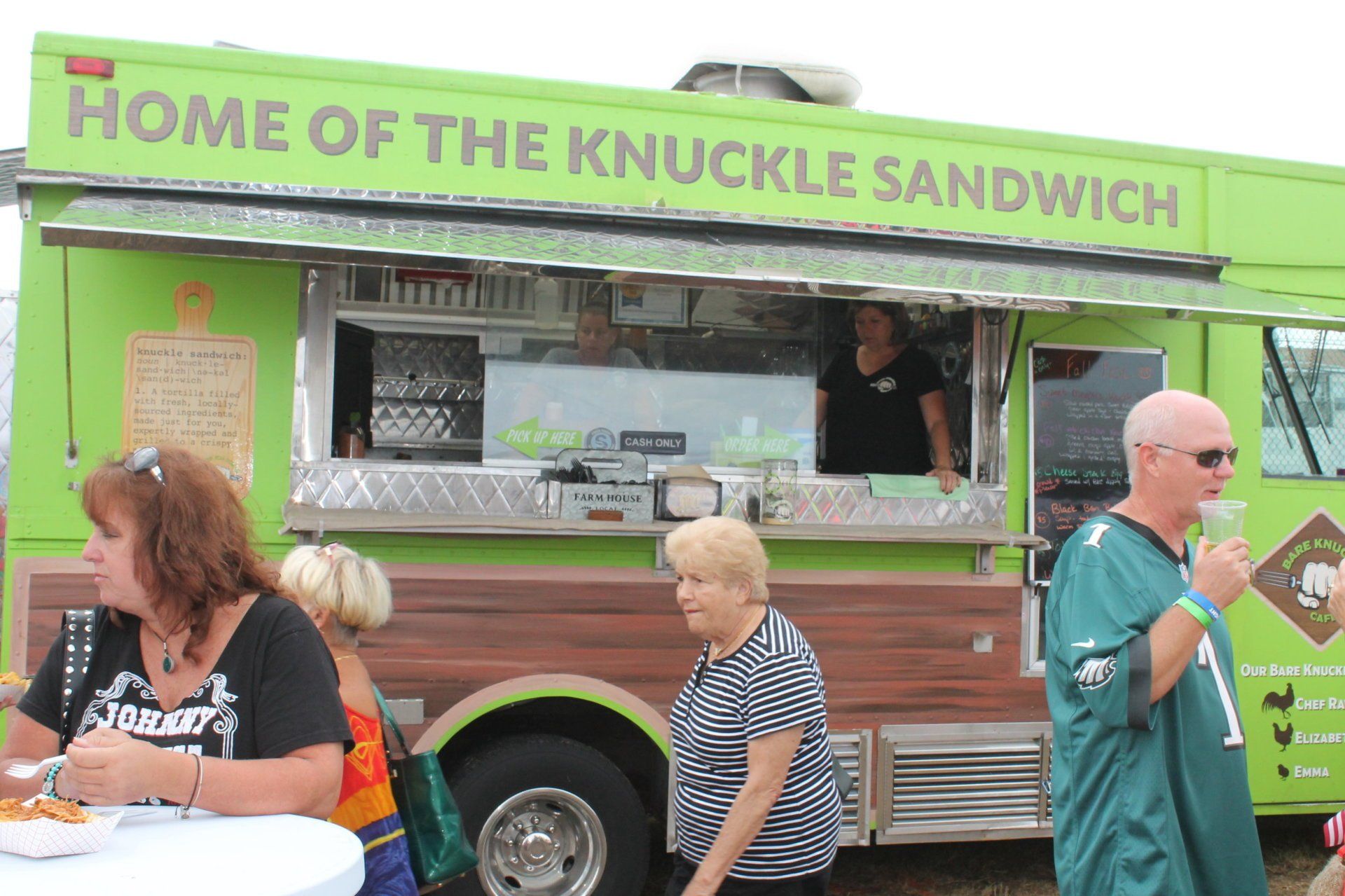 A group of people are standing in front of a green food truck that says home of the knuckle sandwich.