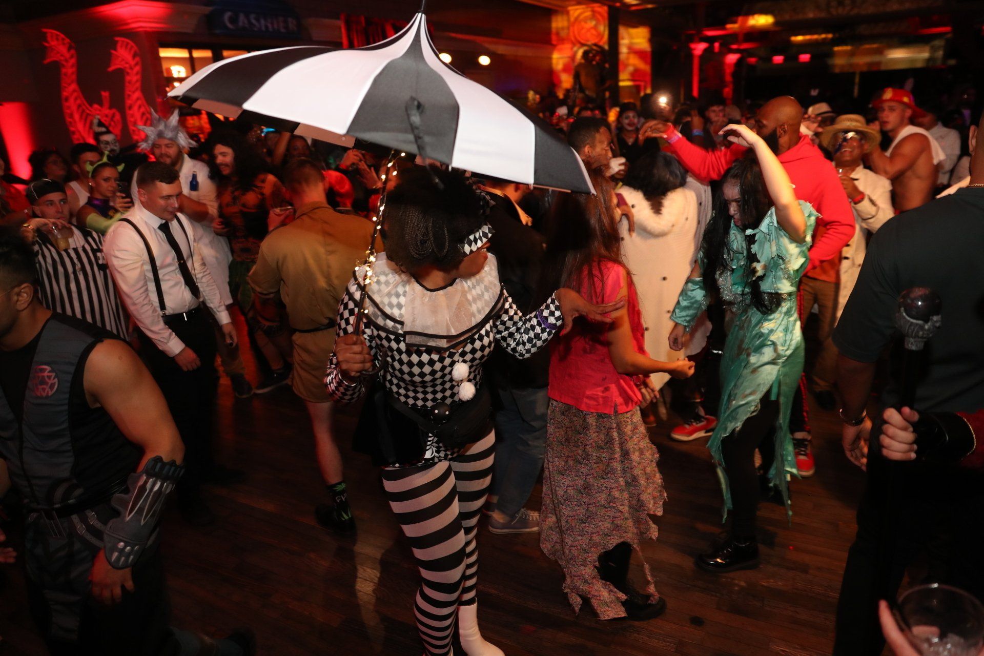 A group of people are dancing in a room with a clown holding an umbrella.