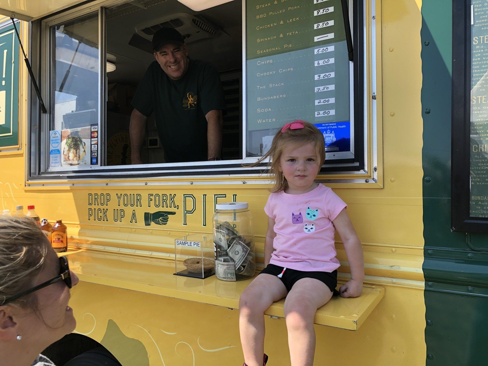 A little girl is sitting on a ledge in front of a food truck.