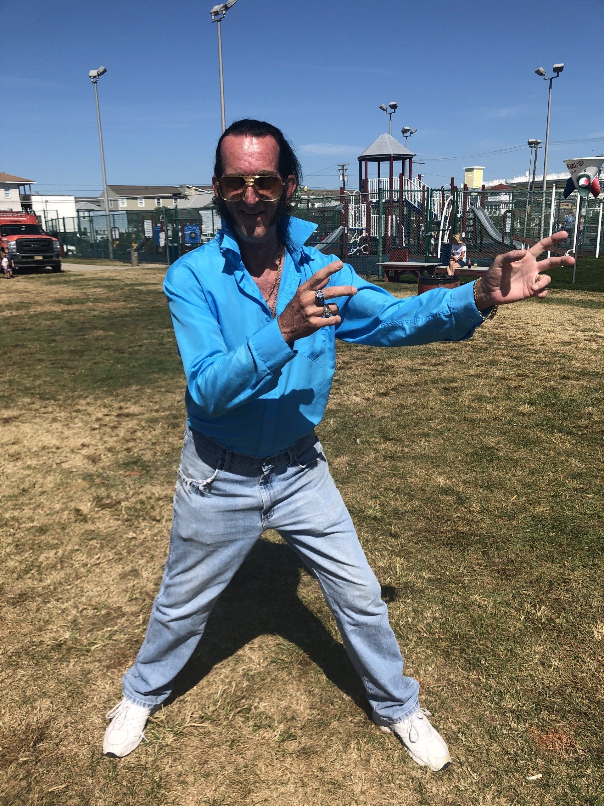 A man in a blue shirt and jeans is standing in a park with his arms outstretched.