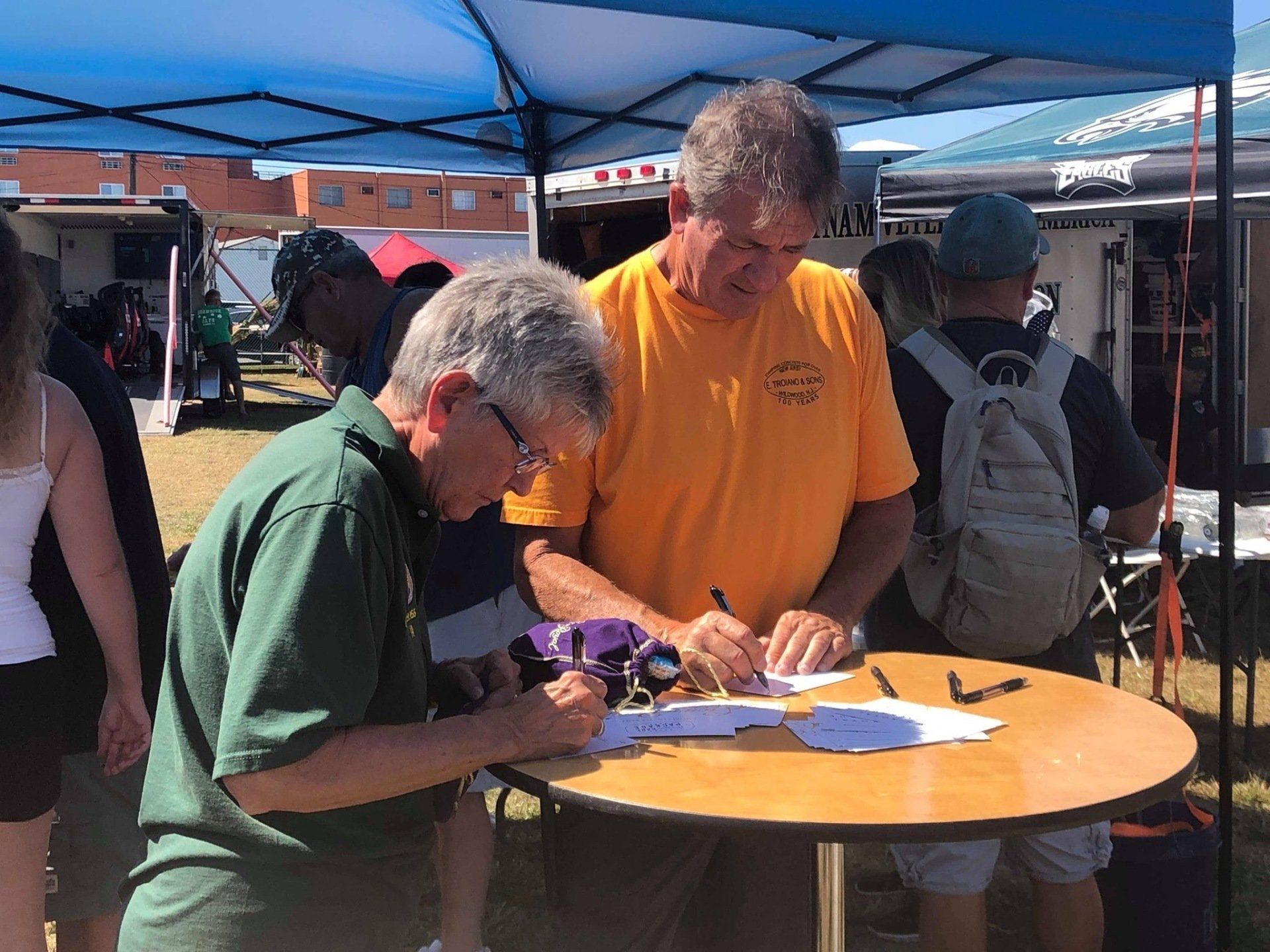 Two men are signing papers at a table under a tent.