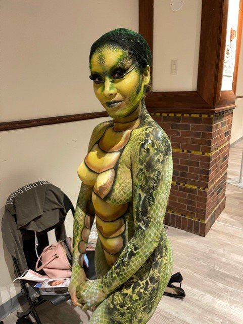 A woman with her body painted like a snake is standing in front of a brick wall.